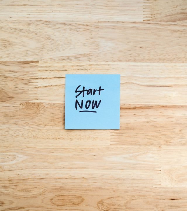 Blue Post-It note with 'Start Now' on light wooden background, inspiring action.